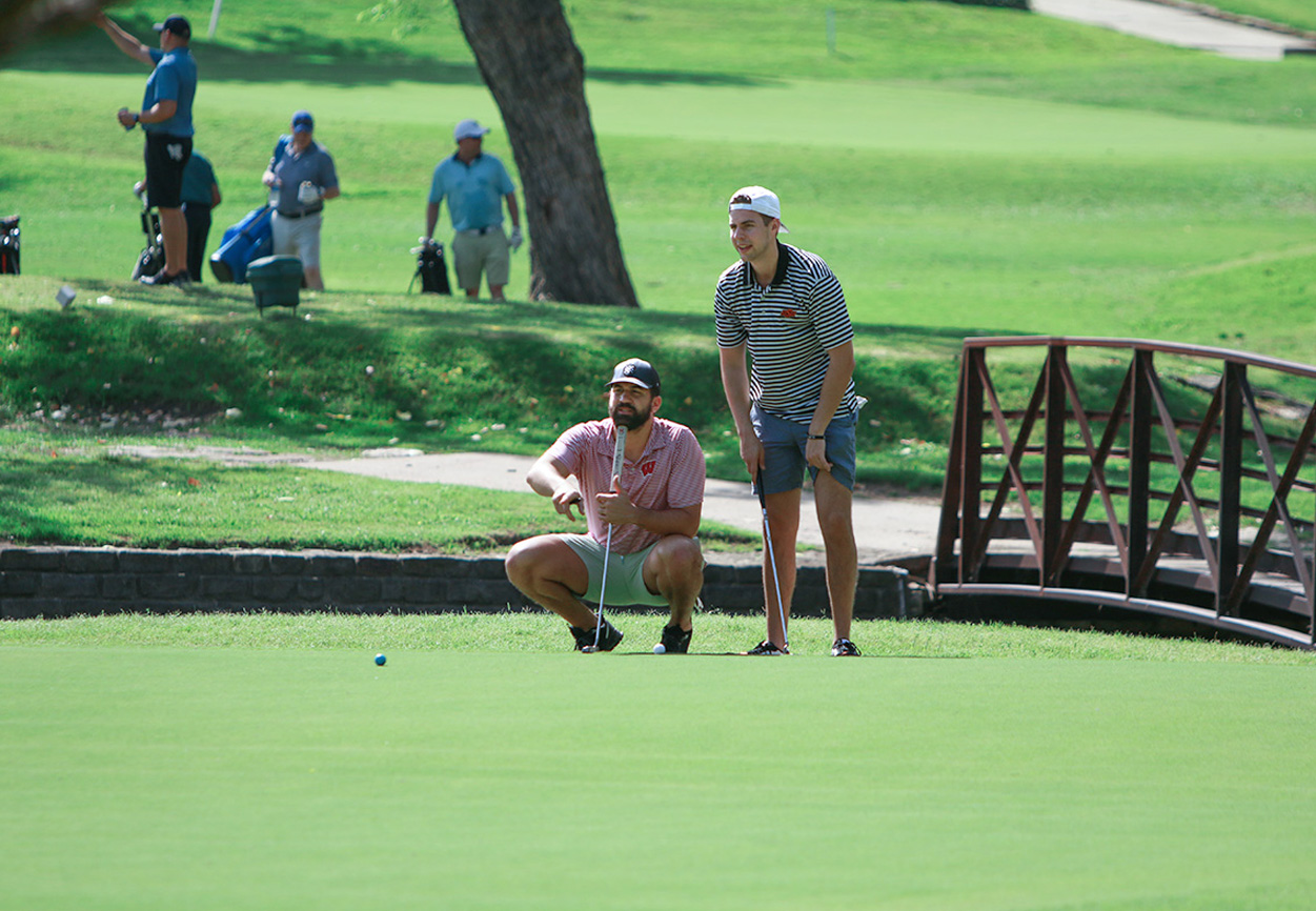 Two golfers on the green during a charity golf tournament, with other participants and a bridge in the background.